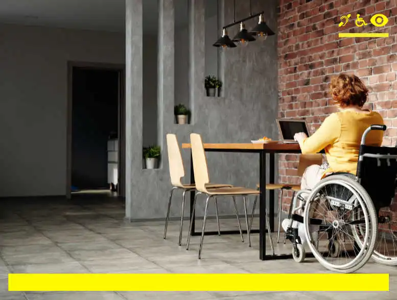 Osoba niepełnosprawna znajdująca się na wózku inwalidzkim pracująca na laptopie. Osoba znajduje się w pomieszczeniu. Ściany pomieszczenia wyłożone są czerwonymi cegłami.